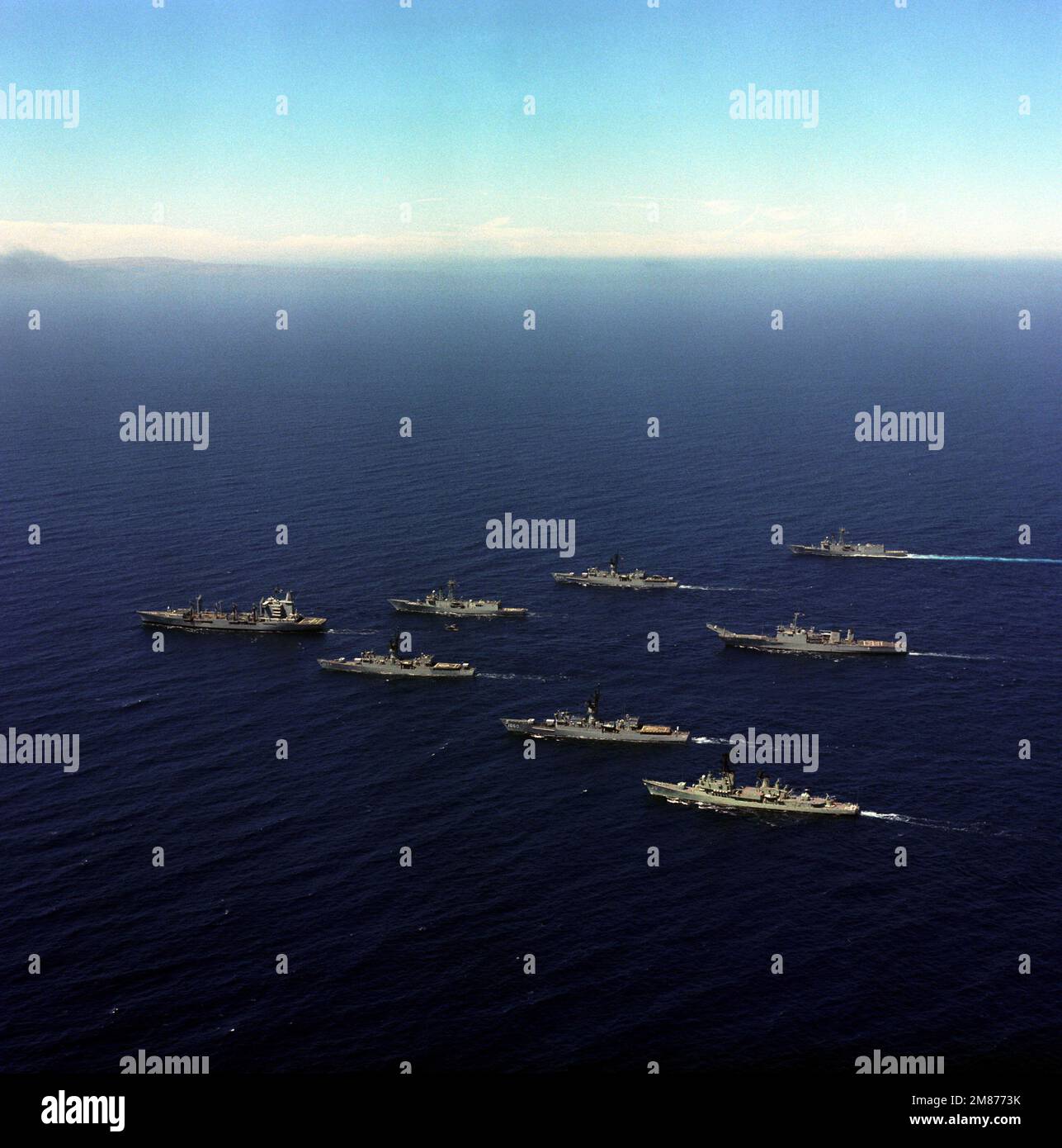 An aerial view of a US Navy (USN) parade formation underway. The ships include, clockwise starting at lower right: USN Farragut Class Guided Missile Destroyer USS MACDONOUGH (DDG 39), USN Knox Class Frigate USS GRAY (FF 1054), USN Cimarron Class Oiler USS WILLAMETTE (AO 180) (leading) USN Oliver Hazard Perry Class Frigate USS DUNCAN (FFG 10), USN Knox Class Frigate USS STEIN (FF 1065), USN Oliver Hazard Perry Class Frigate USS WADSWORTH (FFG 9) and in center the USN Newport Class Tank Landing Ship USS RACINE (LST 1191). In flight is a SH-2F Seasprite helicopter. (Exact Date Shot Unknown). Coun Stock Photo