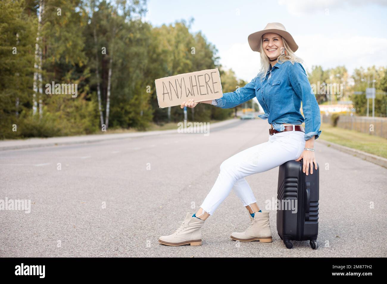 Smiling laughing carefree blond woman in hat and modern jeans shirt hitchhike by roadway with carton nameplate anywhere Stock Photo