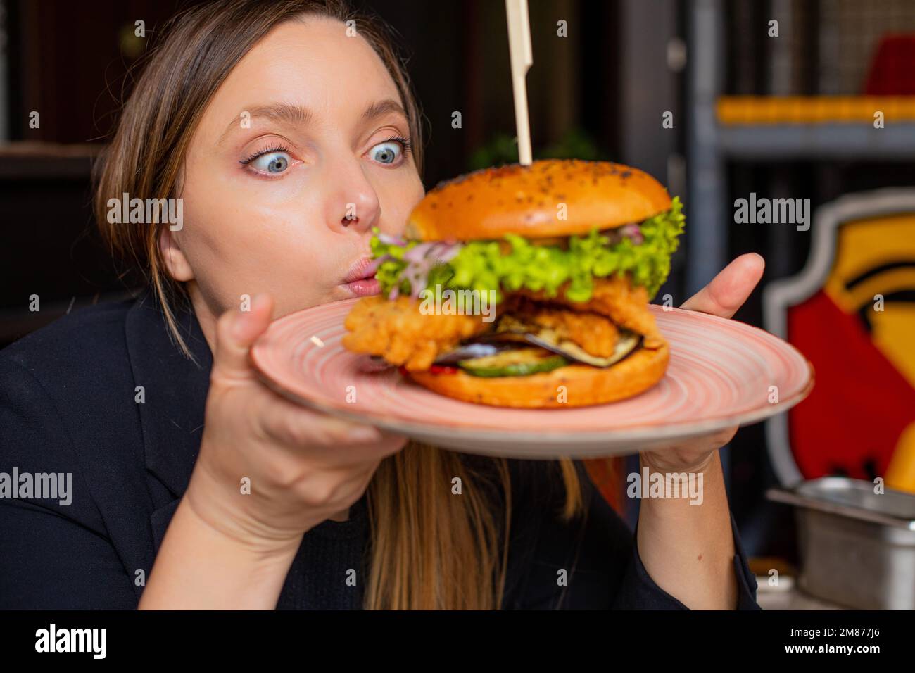 Portrait of surprised woman staring wide-eyed at burger with lettuce, chicken, onion on big plate in cafe restaurant. Stock Photo