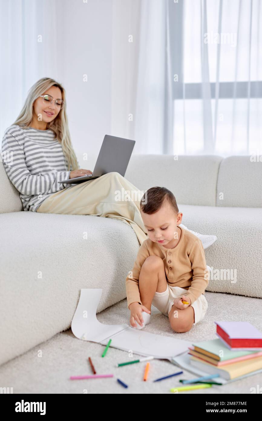Little boy and mom spending weekends at home together, child drawing painting on floor while mother working on laptop in the background. focus on blon Stock Photo