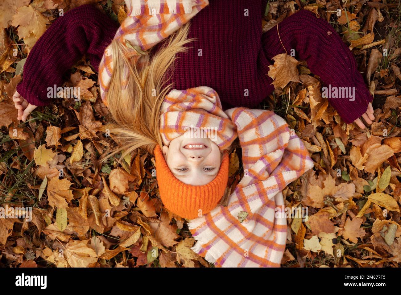 Glad satisfied, delightful little blond girl in warm outfit, scarf and hat play with leaves, lying on maple, oak foliage Stock Photo