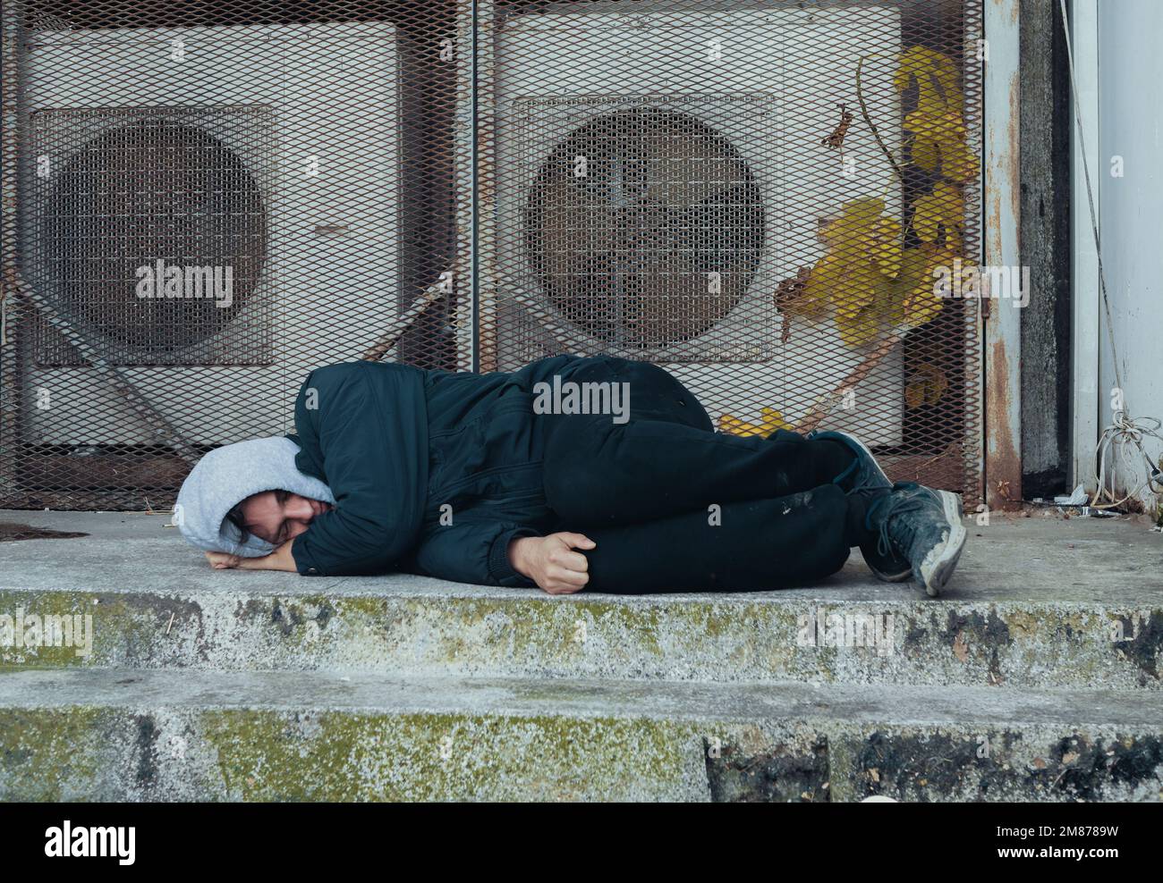 Frozen homeless man sleeps on concrete near building's ventilation system. Problem of how homeless survive winter cold. Stock Photo