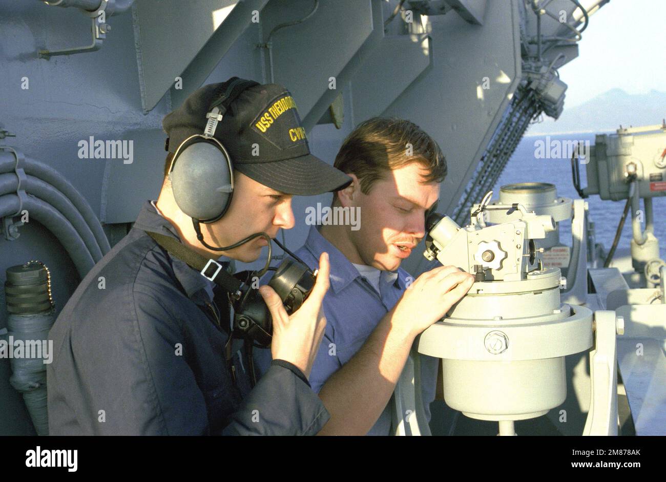 A sailor aboard the nuclear-powered aircraft carrier USS THEODORE ROOSEVELT (CVN-71) uses a telescopic alidade to take a bearing as a phone talker wearing a sound-powered phone stands by to relay the information to the bridge. Country: Unknown Stock Photo