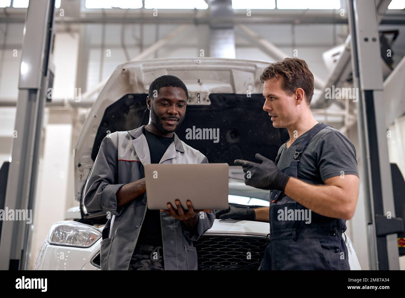 Black Car Mechanic And Caucasian Repairman Checking Diagnostics Results on Laptop, Computer. Explains an Engine Breakdown to Mechanic. Car Service Emp Stock Photo