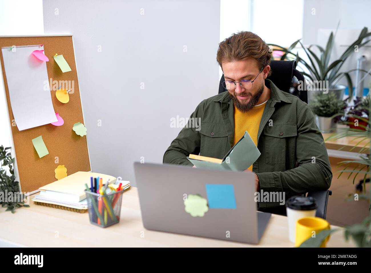 Smiling handsome office worker man sit reading plan, holding notebook in hands, bearded caucasian male in casual wear concentrated focused on work, ge Stock Photo