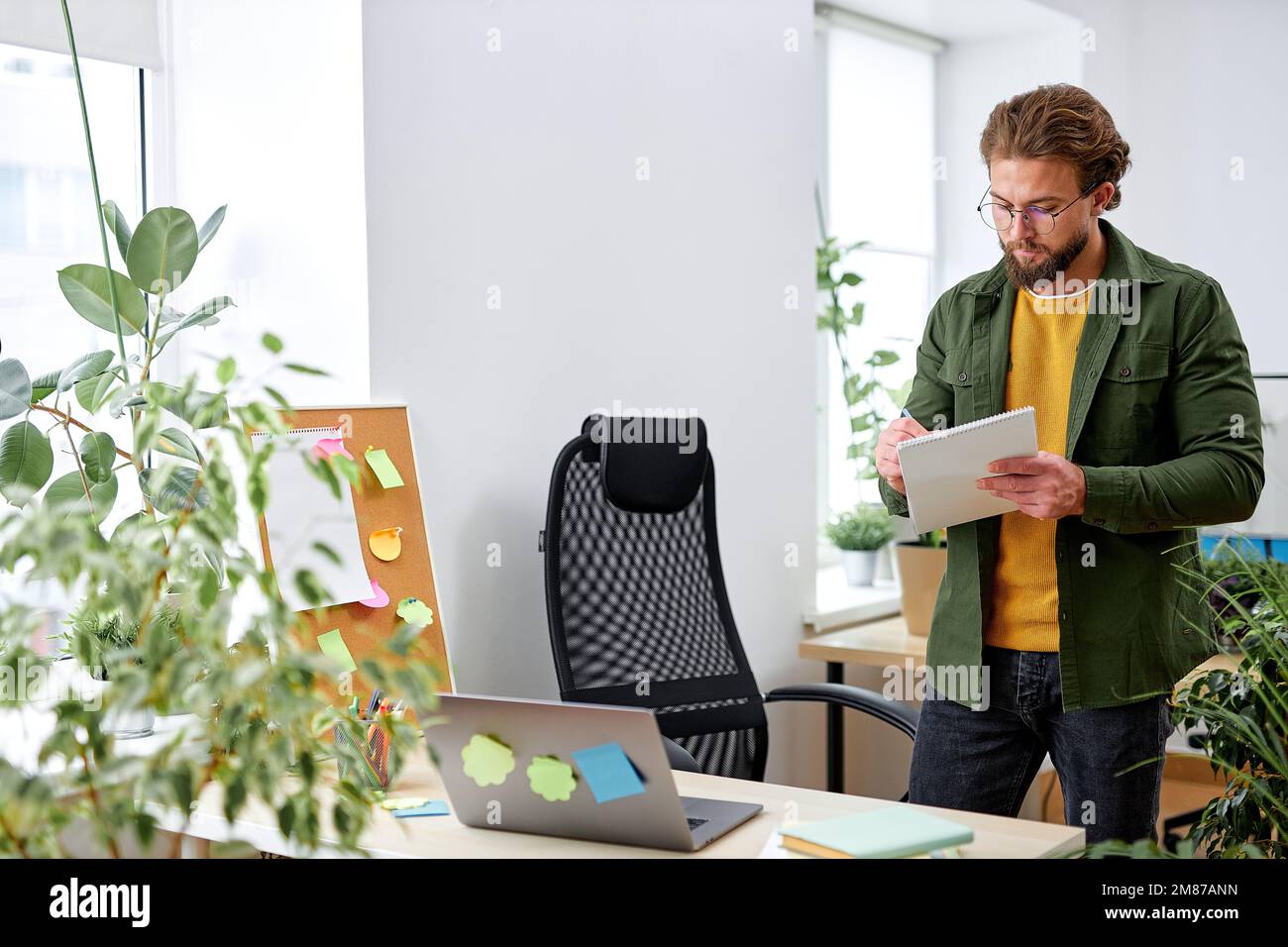 Young man office worker concentrated on work, writing planning, standing behind desk in bright cozy modern office, use laptop, dressed in casual cloth Stock Photo