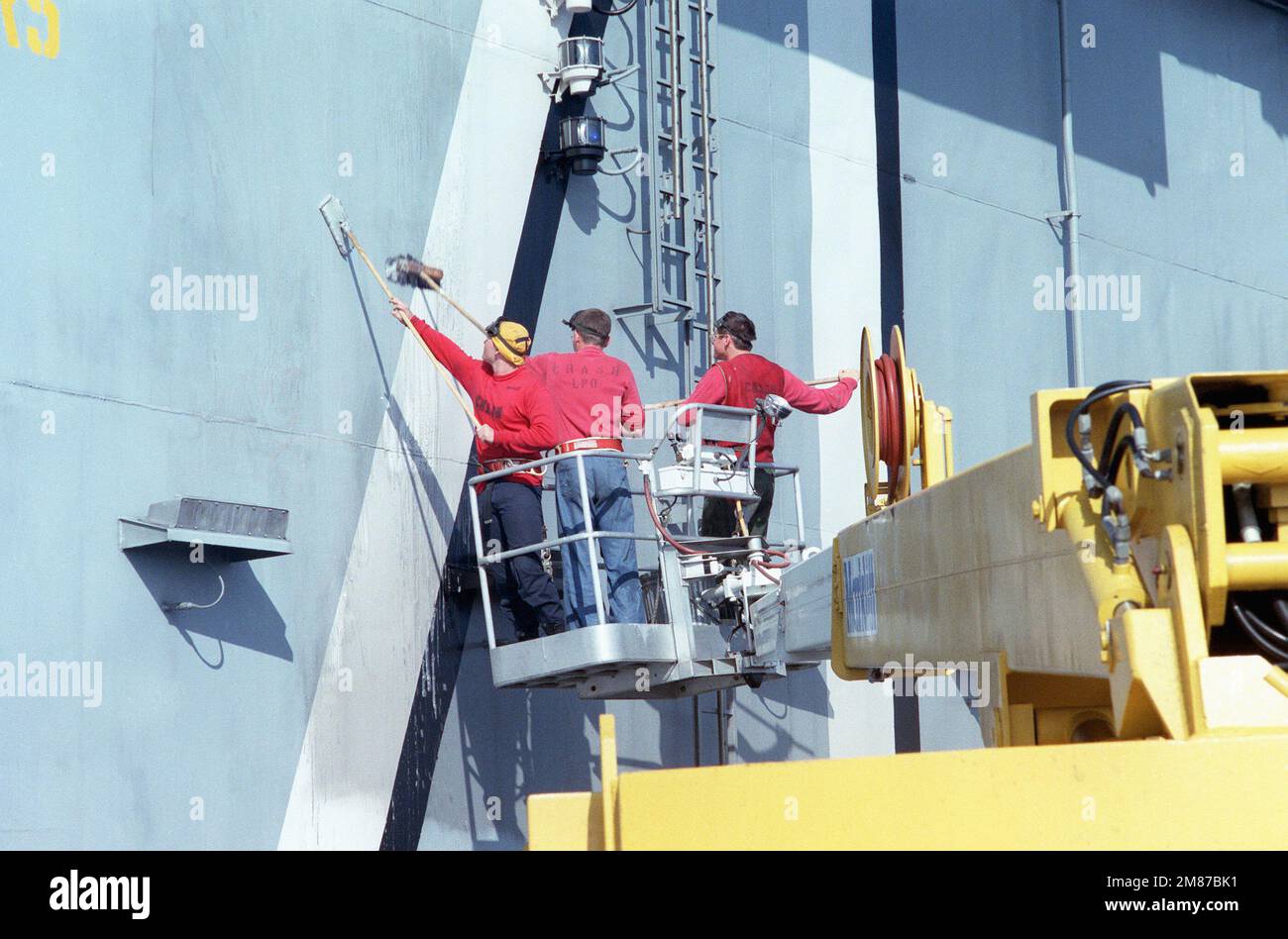 Crewmen stand in a basket at the end of a crane to clean a side of the ...