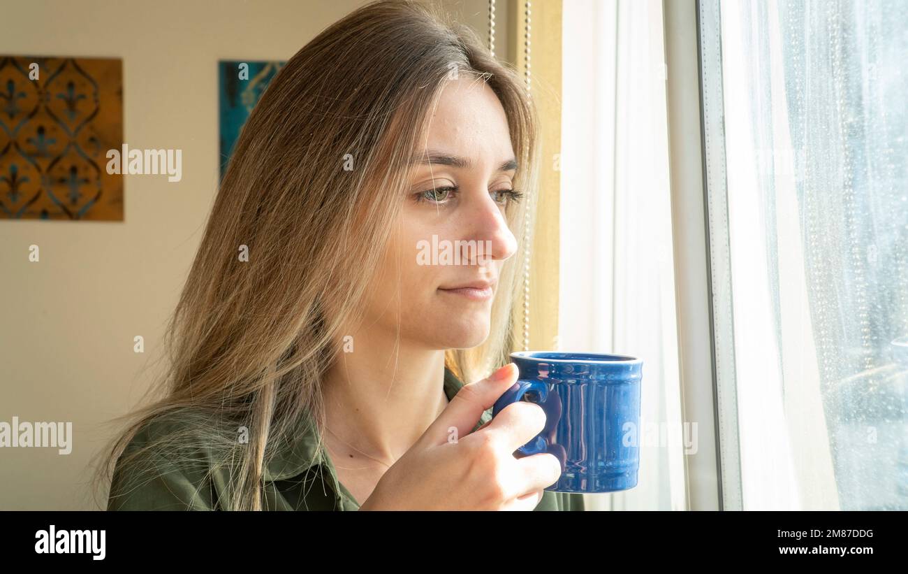 Depressed woman drinking coffee by the window at home. Thoughtful woman questioning life. Young adult girl watching outside Stock Photo