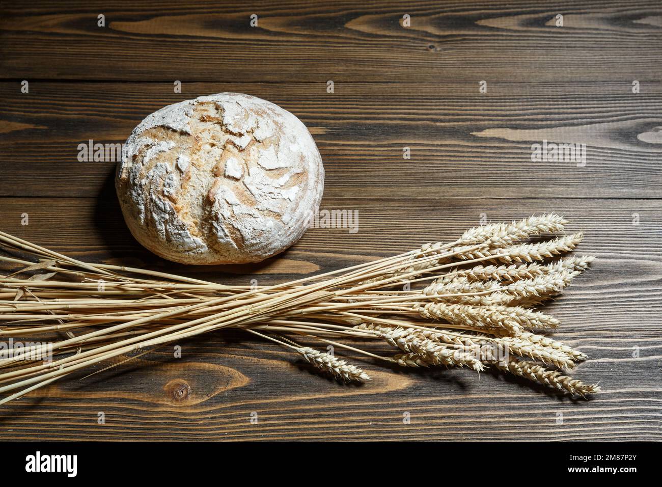 Fresh baked bread loaf and wheat ears lying on a wooden background. Food provision and production concept. Stock Photo