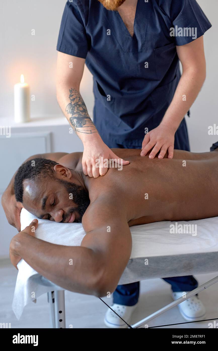 cropped caucasian male masseur with strong hands professionally massaging scapulas and shoulders of male. handsome naked young black man with perfect Stock Photo