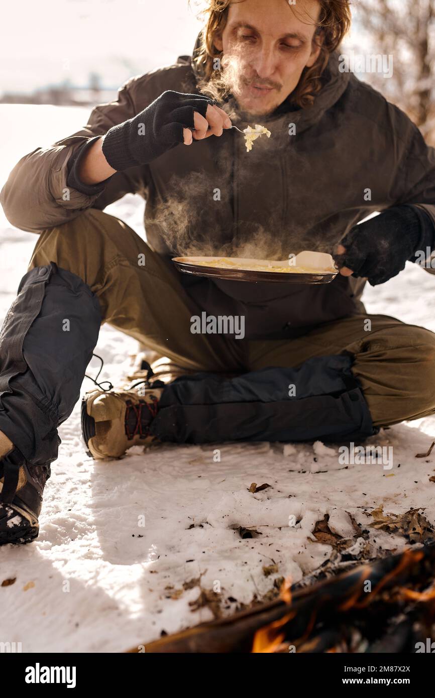 young caucasian man traveler cook scrambled eggs on fire. bushcraft, adventure, travel, tourism, hike and camping concept. handsome guy in warm coat s Stock Photo