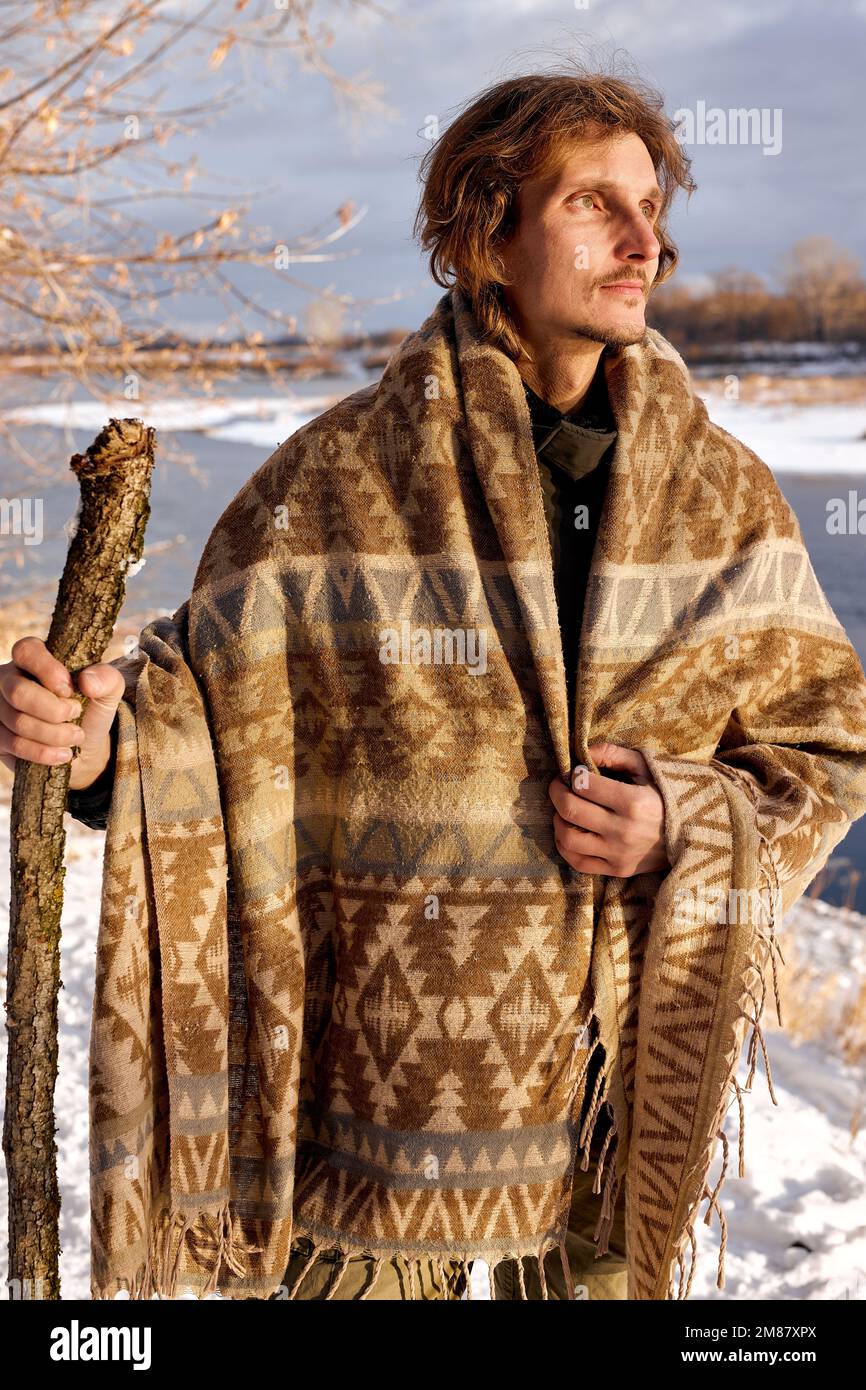Young tourist with wooden stick looking around while walking in winter forest on cold day, wrapped in scarf, at sunny frozen day or morning, bushcraft Stock Photo