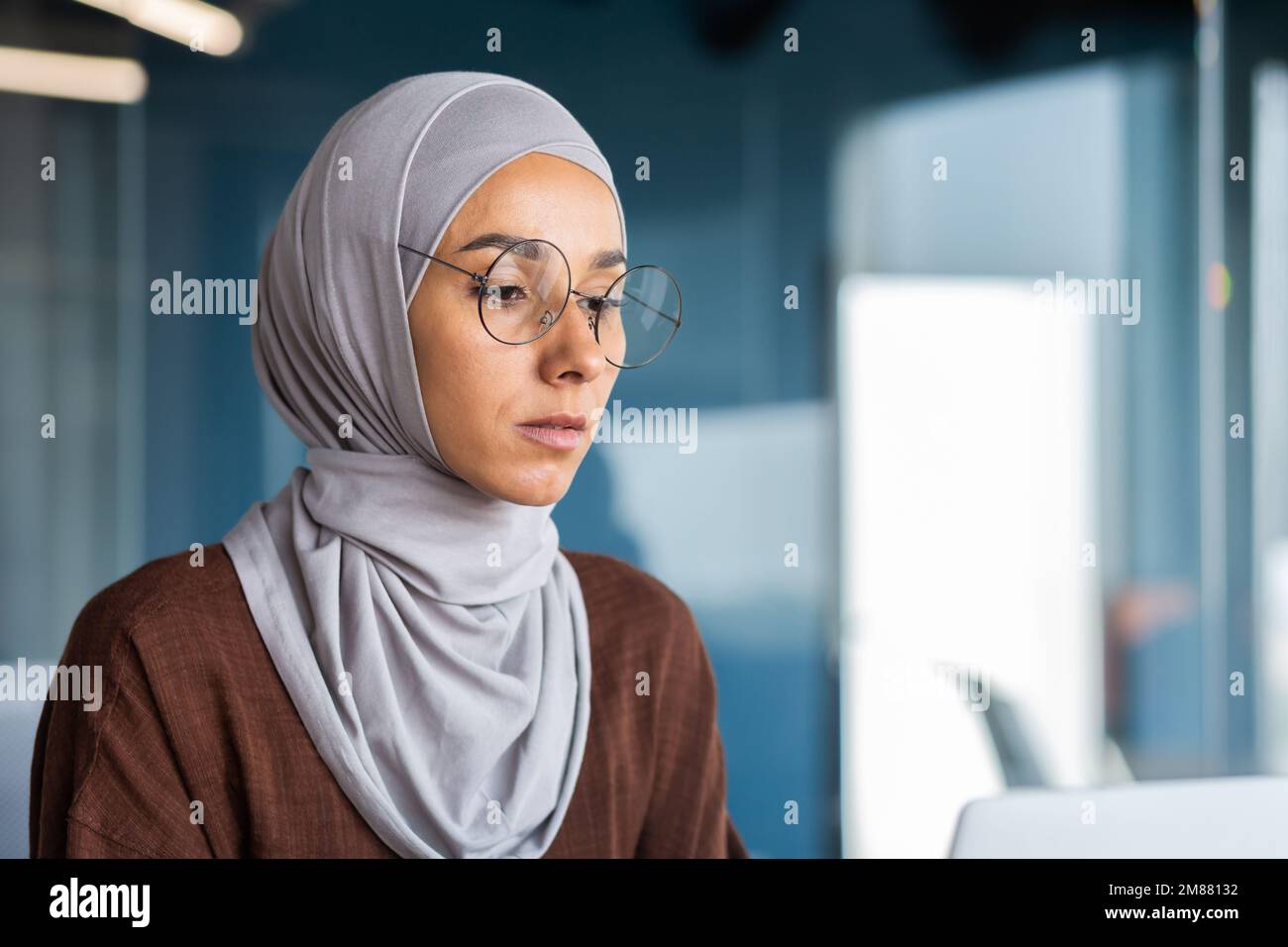 Close up of thinking business woman in hijab in modern office, serious muslim woman looking at laptop screen thinking about decision, woman at workplace successful. Stock Photo
