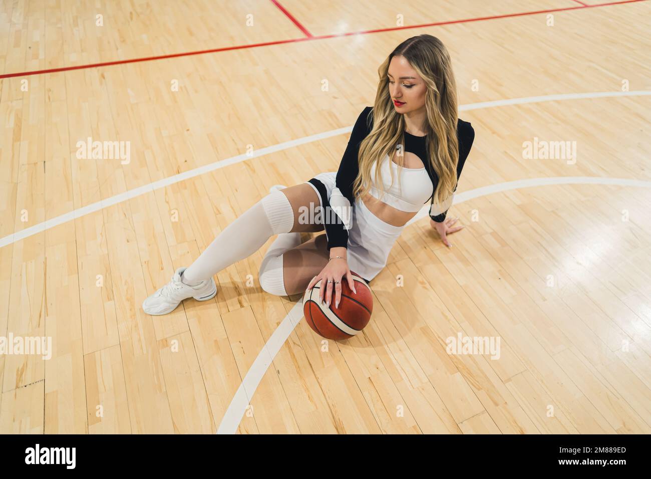 An attractive cheerleader with blond hair sitting confidently on the arena floor with a basketball. Dressed in a mini-skirt and knee-high socks. Showing sports spirit. Stock Photo