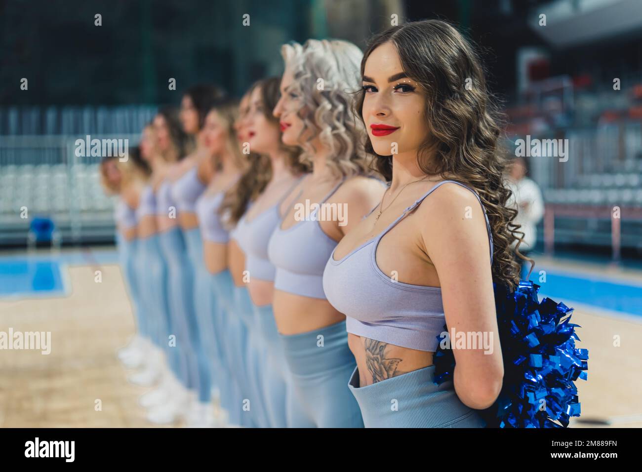 A cheerleader stands confidently in a row with her fellows. Blue pom-poms are held in their hands. The cheerleader's warm smile radiate positivity. Perfectly capturing the spirit of cheerleading. Stock Photo
