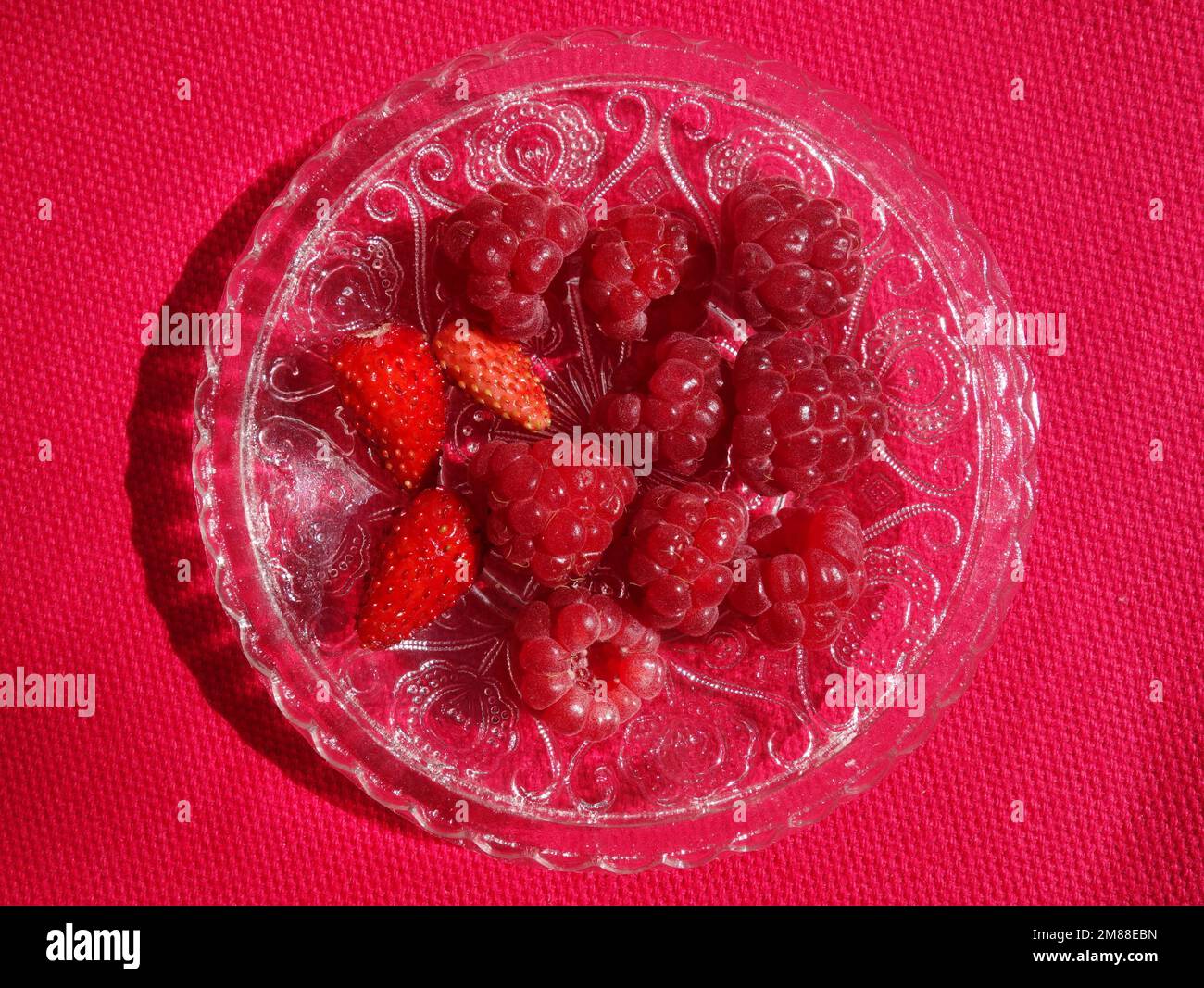 resh raspberries and strawberries fruits healthy snack. Heap of red berries on ornamented glass saucer on magenta cloth Stock Photo