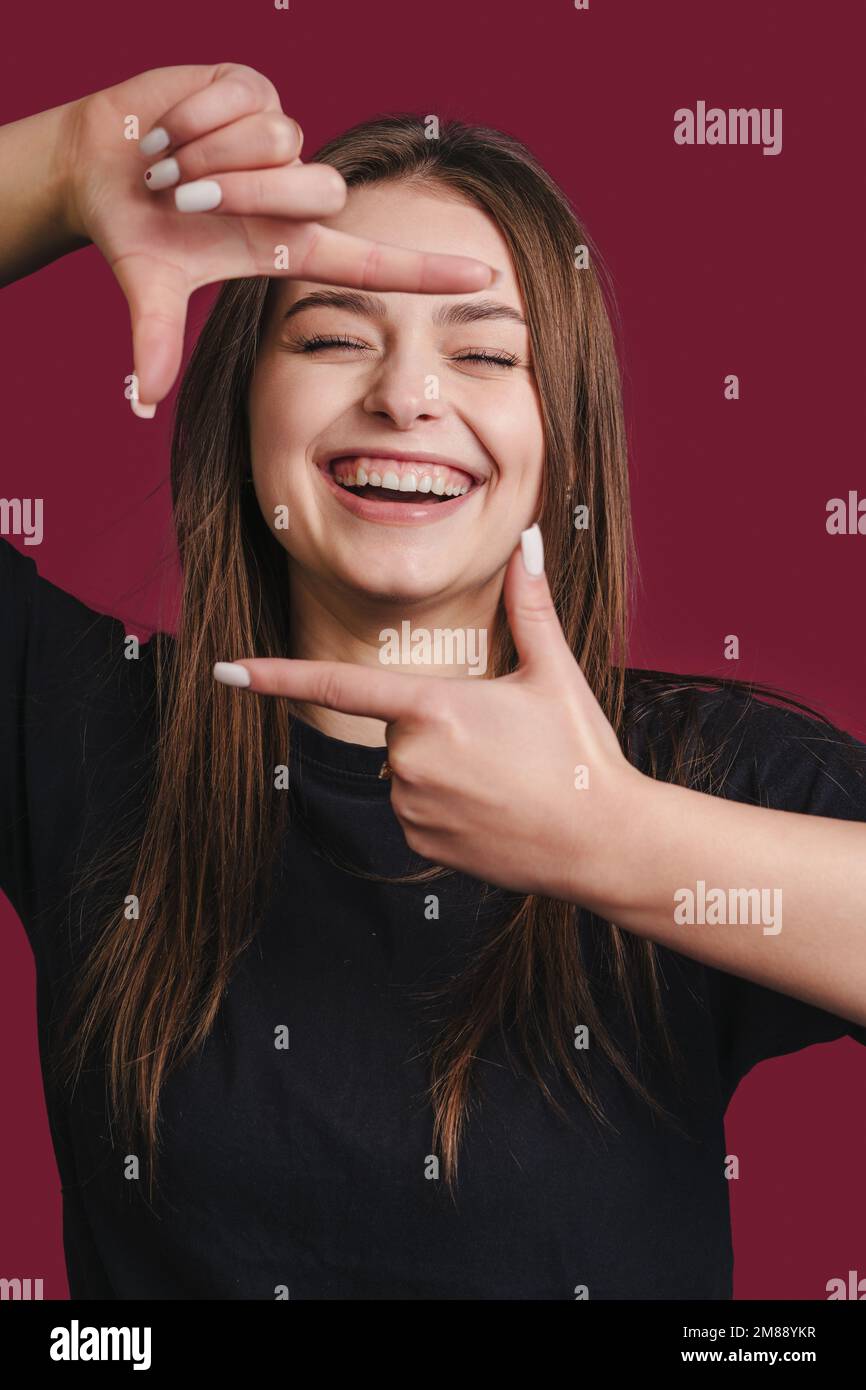 Portrait of young beautiful woman cheerfuly smiling making a camera frame with fingers isolated over cherry-colored background. Stock Photo