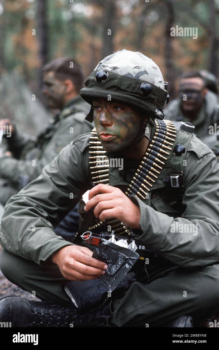Security Police student, in camouflage face paint and MILES equipped helmet and wearing a M-60 machine gun ammo (ammunition) belt around his neck, takes a break of MRE's (Meals Ready to Eat). He is receiving Army Base Ground defense training during Army conducted course in combat skills, at Fort Dix, New Jersey. Exact Date Shot Unknown. Country: Unknown Stock Photo