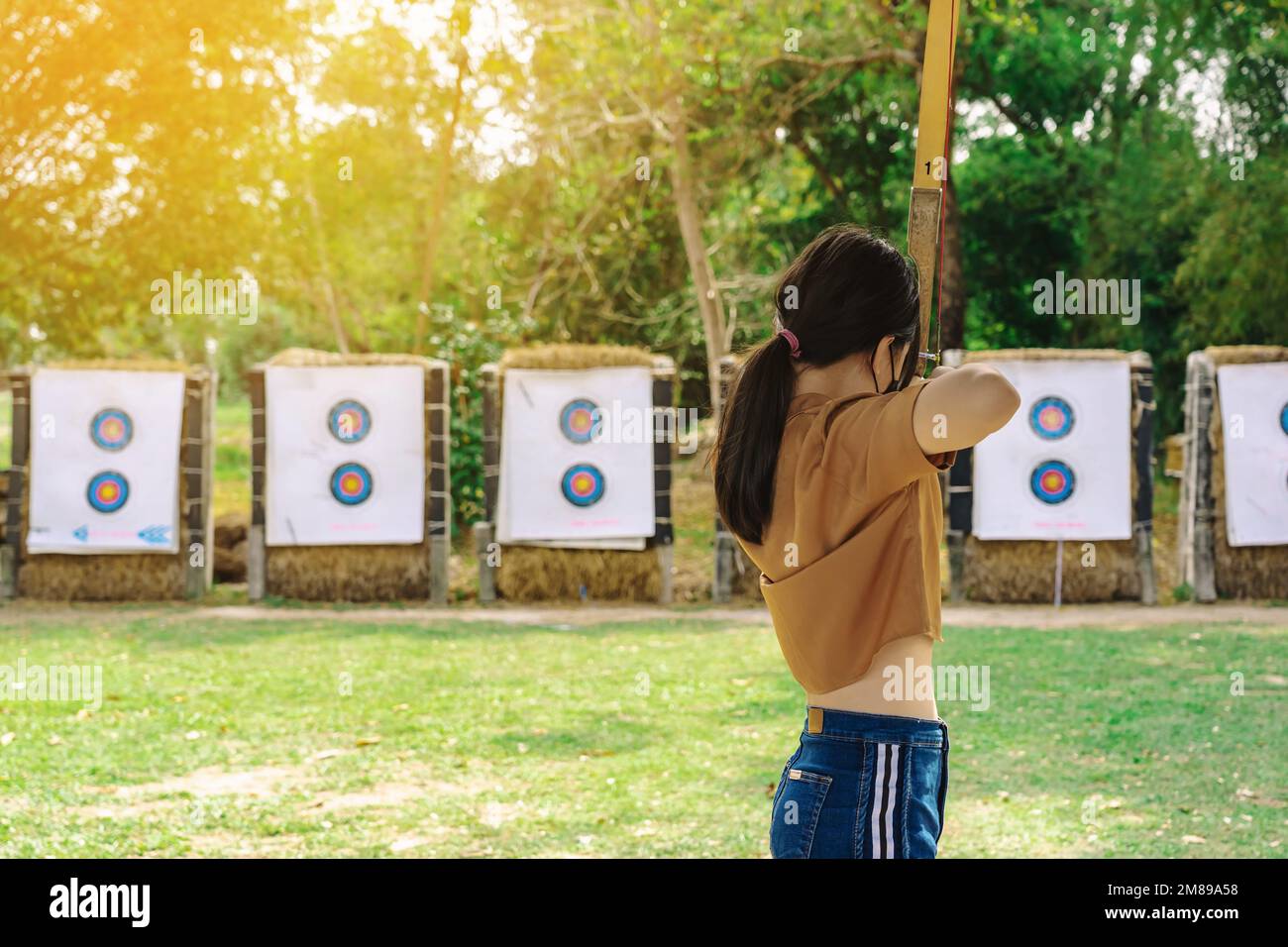 Back view of Asian girl wear face mask aims archery bow and arrow to colorful target in shooting range during training and competition. Exercise and c Stock Photo