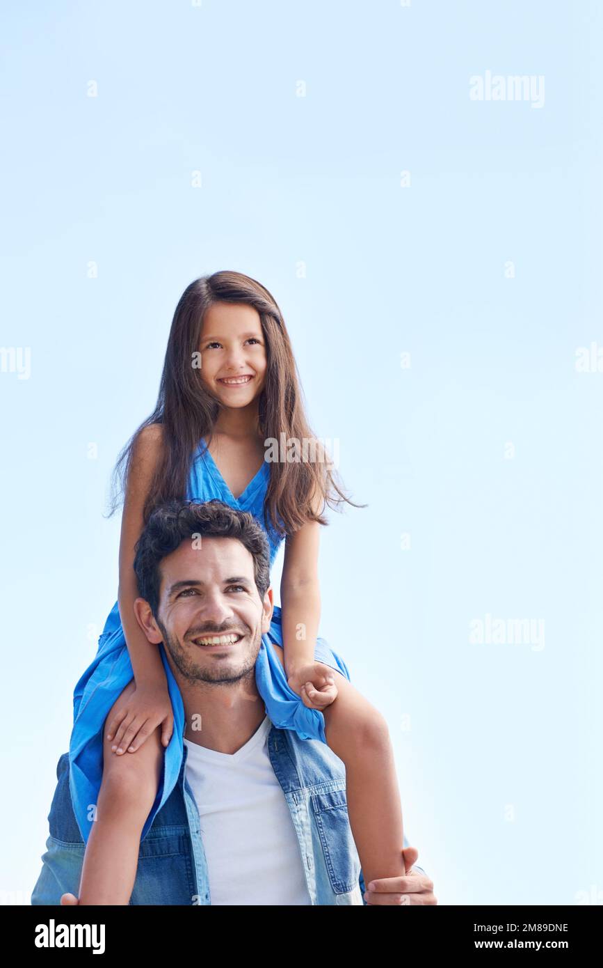 When Im with Daddy, Im on top of the world. Low angle shot of a girl sitting on her fathers shoulders against a blue sky. Stock Photo