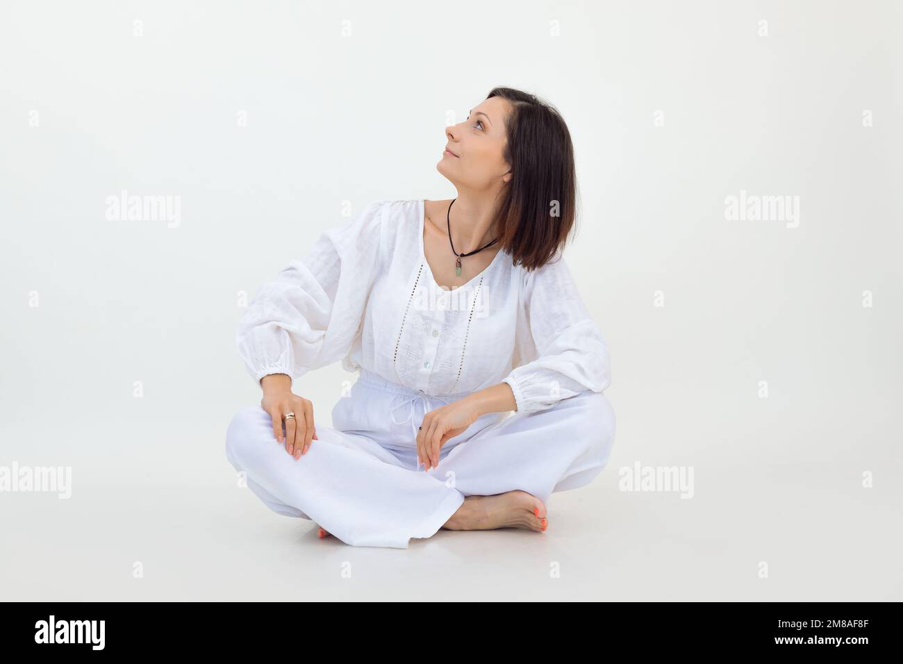 Young woman wearing white loose blouse, trousers, sitting on floor with crossed legs, looking up on white background. Stock Photo
