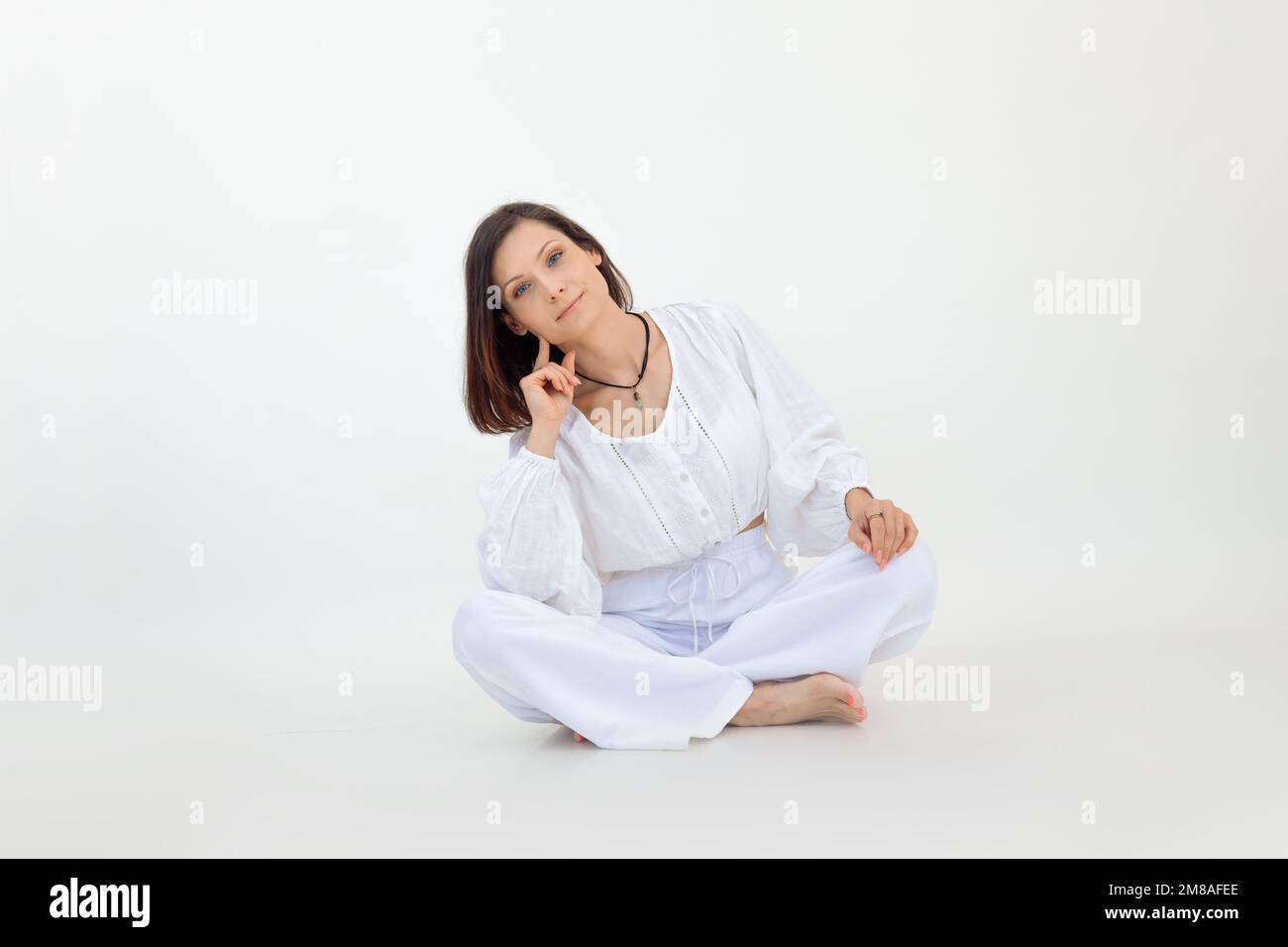 Portrait of young woman with short dark hair wearing white loose blouse, trousers, sitting on floor with crossed legs. Stock Photo