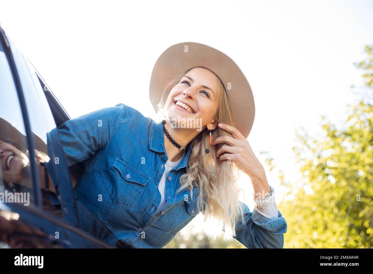 Laughing merry glad pensive blond woman in hat, jeans shirt lean out of car, joy fresh air. Carpool, rental for holiday Stock Photo