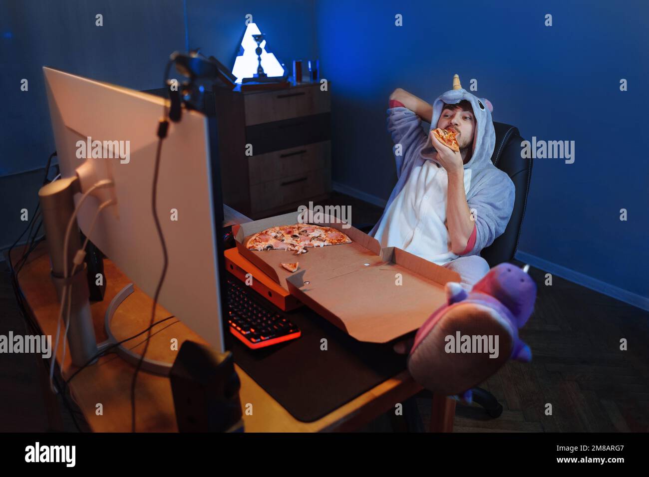 Young funny man in unicorn costume watching live stream of video game tournament on professional gaming computer, eating pizza, having fun. Guy enjoying series, TV show, resting at home. Leisure time Stock Photo
