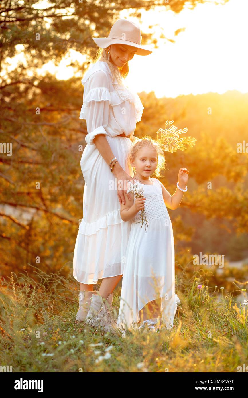 Happy mother with cute little daughter stand in white dresses in wood at yellow sunset, sun shining. Mom and child walk in forest at summer evening Stock Photo