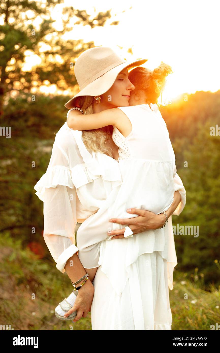 Portrait of beautiful lady in hat with love holding in hands little girl outdoors in wood at sunset. Mom and child walk in forest at summer evening Stock Photo