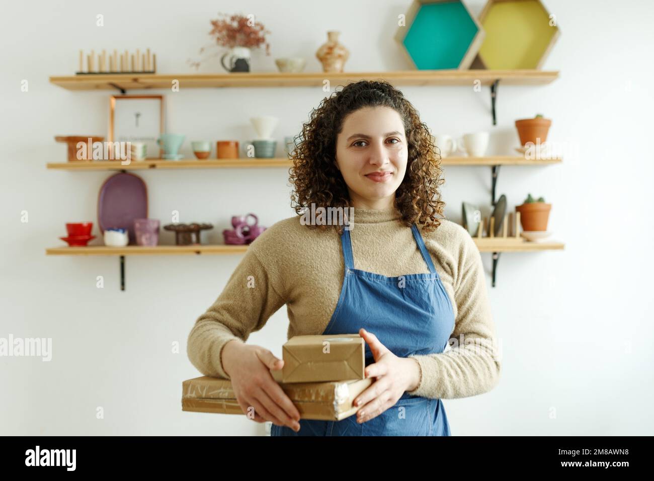 Vertical waist up portrait of young female artisan looking at camera while standing with boxes in workshop, hobby and small business concept. Stock Photo