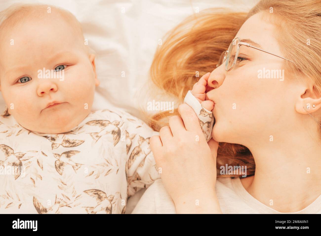 Cheerful young mother kiss fingers on palm of little baby lying on bed closeup. Home family photo of woman and cute infant child resting together Stock Photo