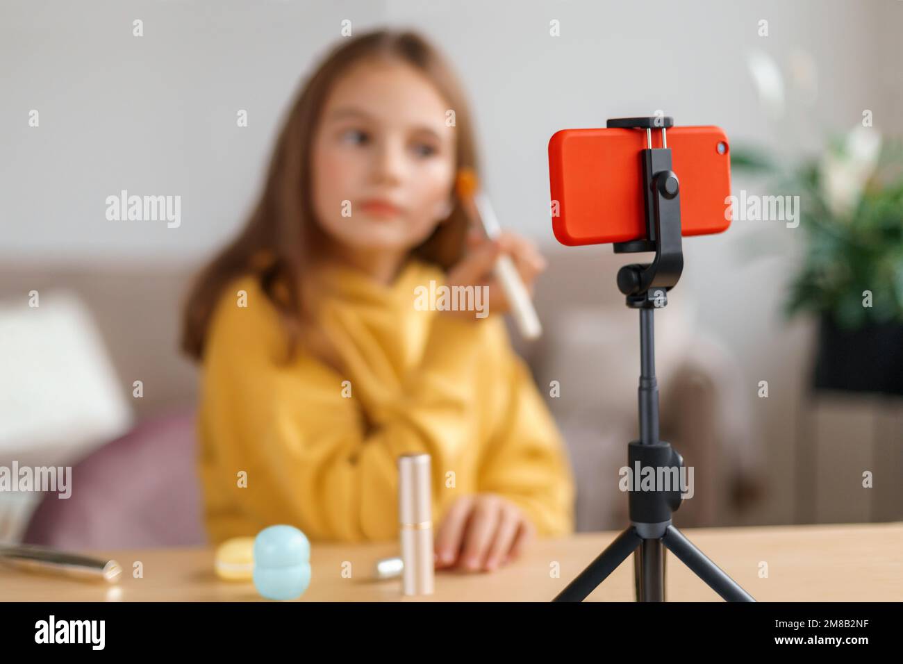 Young beautiful girl doing makeup with kid cosmetics on smartphone camera while shooting online tutorial for her followers. Cute little beauty blogger recording vlog at home to post it on social media Stock Photo