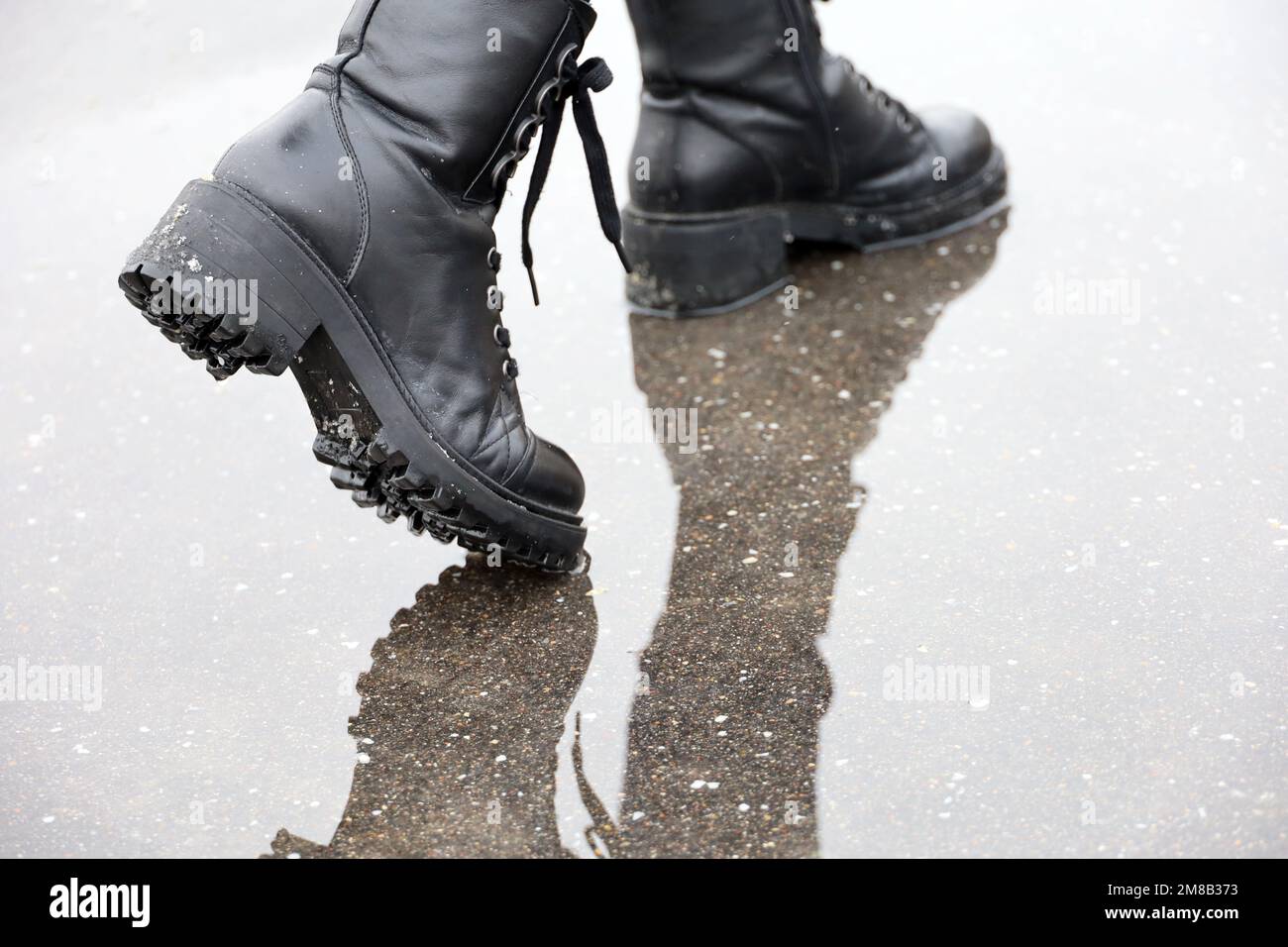 Female legs in black leather boots with reflection in puddle on a street. Rain with snow in city, winter or spring weather, waterproof footwear Stock Photo
