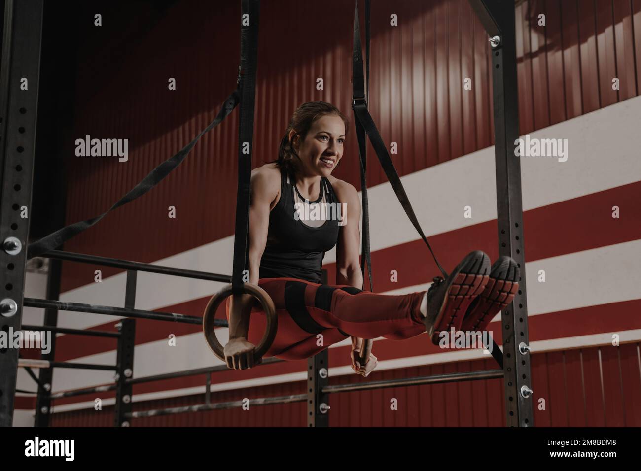 Cheerful crossfit woman doing abs exercises on gymnastic rings. Female athlete works on her abdomenal muscles during workouts at the gym. Stock Photo