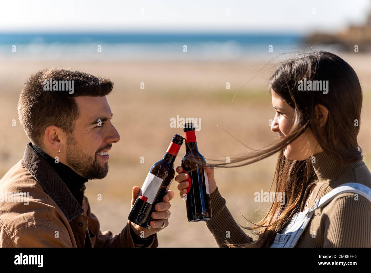 Young boyfriend and girlfriend smiling and clinking bottles of beer during Saint Valentine Day celebration on blurred background of sea and beach Stock Photo