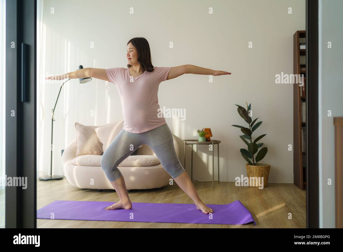 Full length healthy 6 months pregnant calm Asian woman meditating or doing yoga exercise at home. Relaxation and stretching. Stock Photo