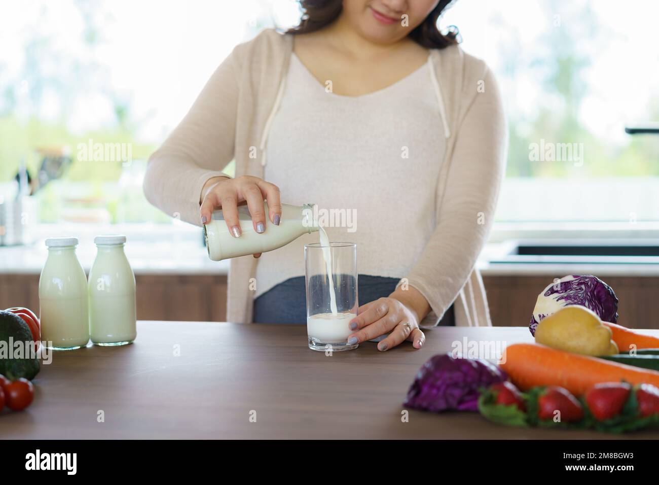 Charming Asian pregnant woman is holding bottle of milk and pouring milk to a glass. Attractive woman need protein and calcium for her baby and it mak Stock Photo