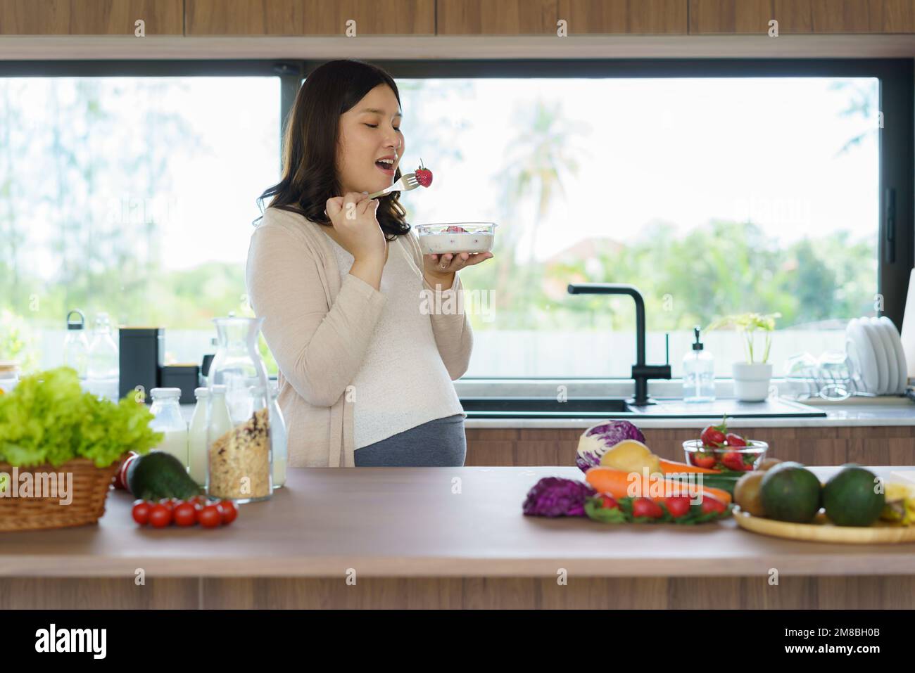 Beautiful Asian pregnant woman eating with lust cereals on breakfast in kitchen at home, enjoying meal. Yummy food for pregnant Stock Photo