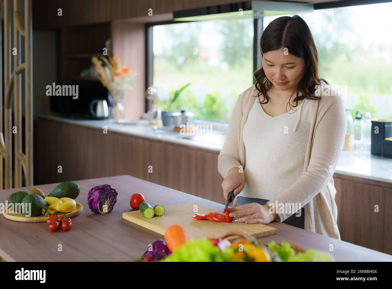 Pregnant Asian woman cutting tomato for fresh green salad, female prepares tasty organic dinner at home, healthy nutrition for future mother Stock Photo