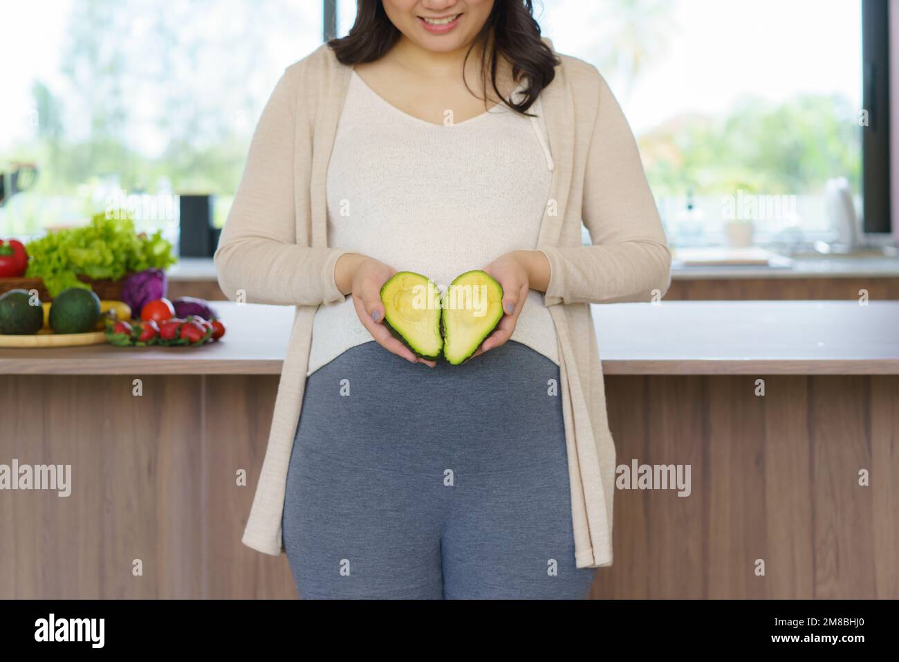 Asian pregnant woman holding fresh avocado fruit close to her belly in kitchen at home. Healthy nutrition and pregnancy concept Stock Photo