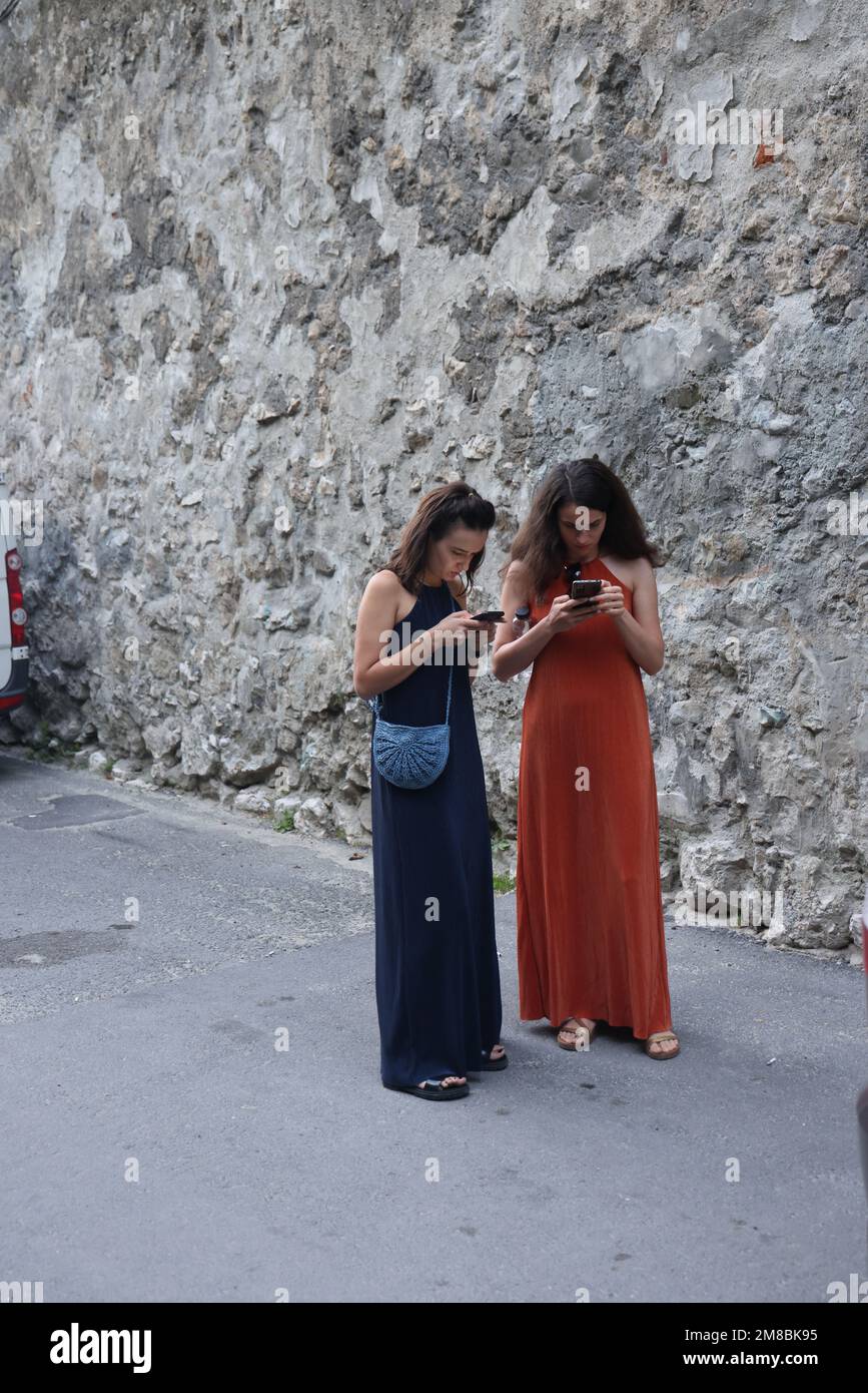 Two females in long dresses and on open shoes looking on their phones, possibly looking for the direction in navigation apps or on maps; conceptual Stock Photo