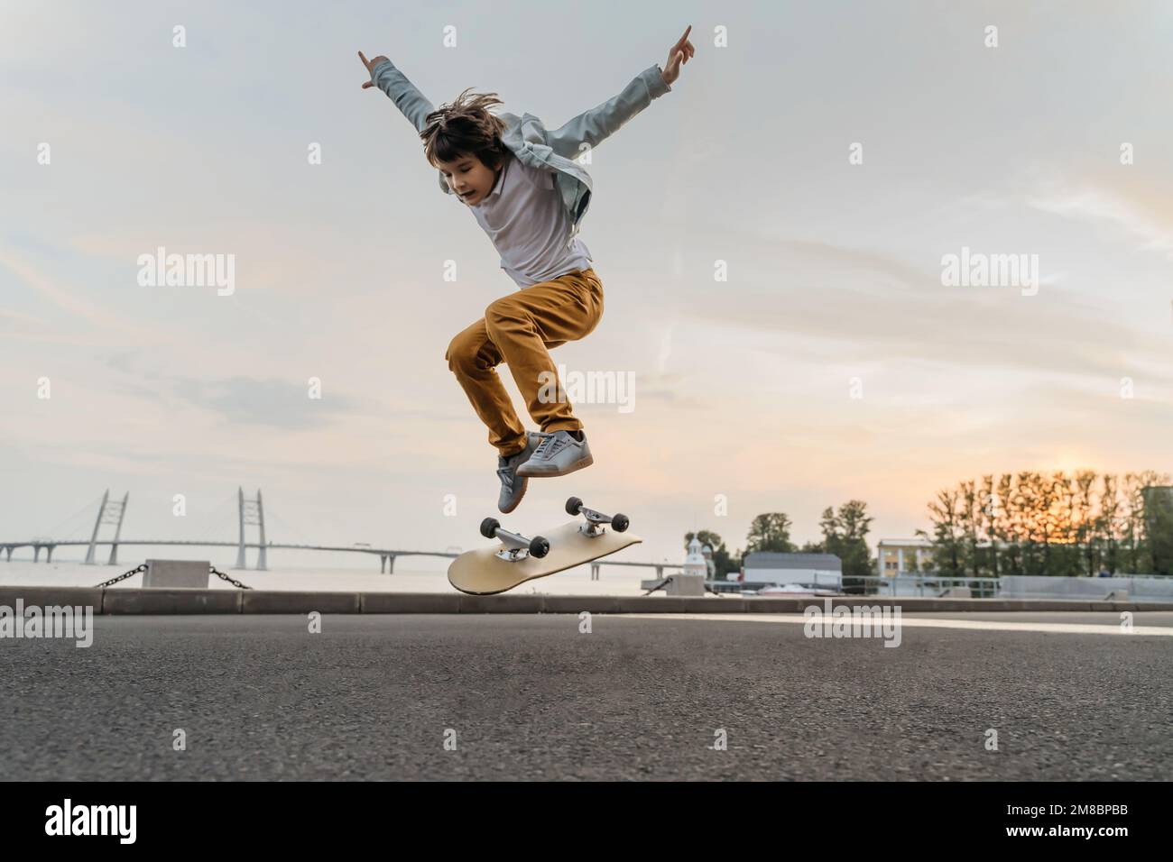 Boy jumping on skateboard at the street. Funny kid skater practicing ollie on skateboard at sunset. Stock Photo