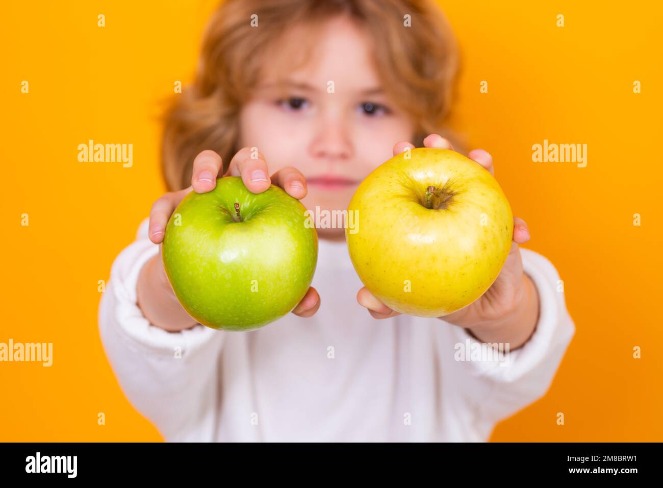 Kids face with fruits. Close up kid with apple in studio, selective focus. Studio portrait of cute child hold apple isolated on yellow background. Stock Photo