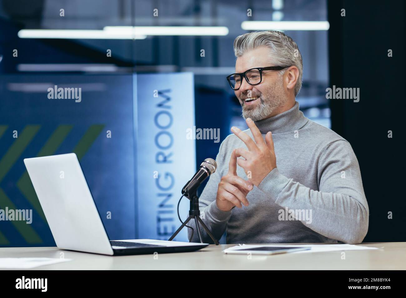 Senior gray-haired man sitting at a desk in an office with a microphone and laptop. Talks on a video call. Conducts interviews, business meetings, webinars, online meetings. Shows, counts on fingers. Stock Photo