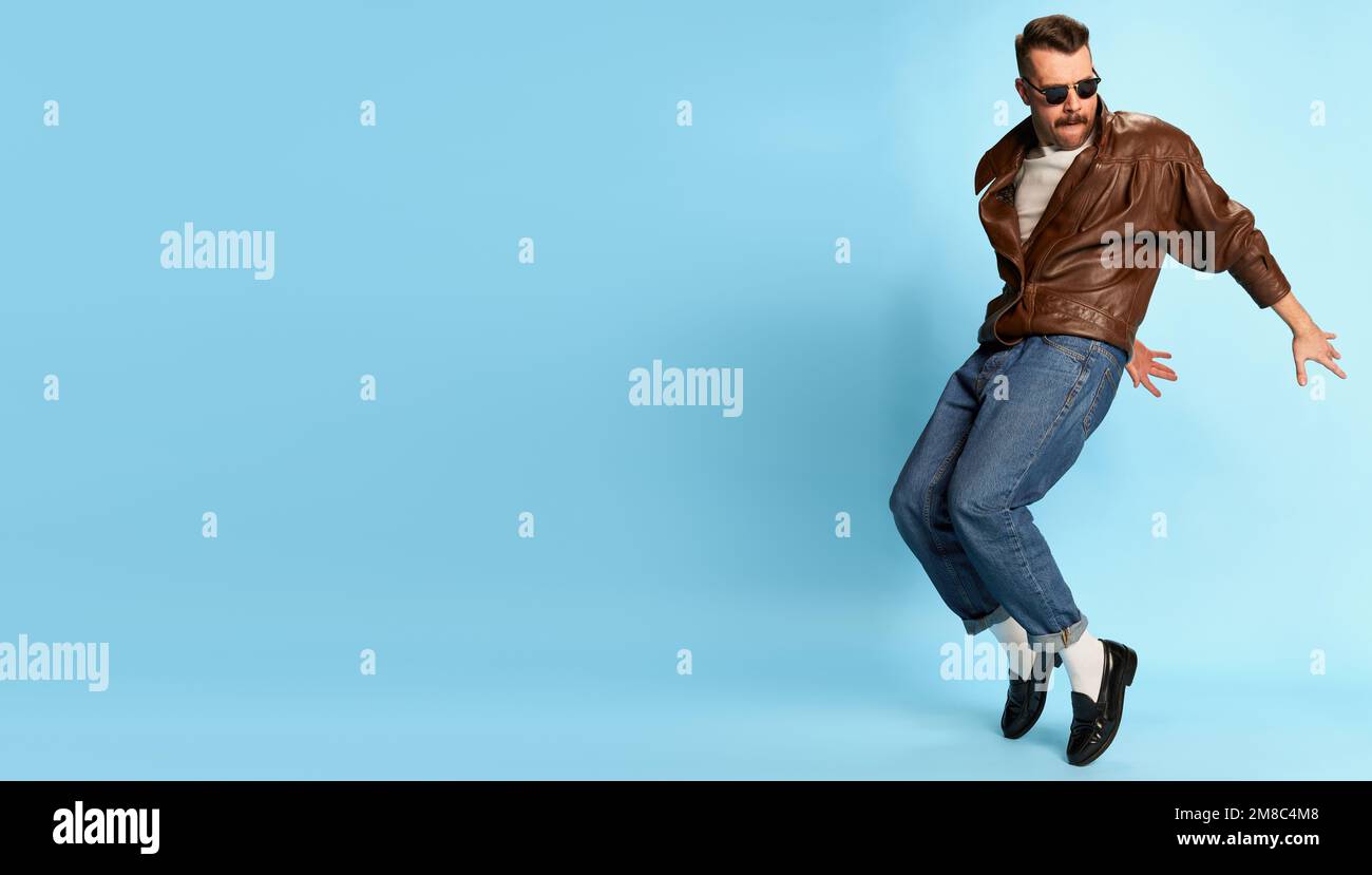 Portrait of brutal, handsome man in jeans, leather jacket and sunglasses posing, dancing over blue studio background. Banner, flyer. Concept of Stock Photo