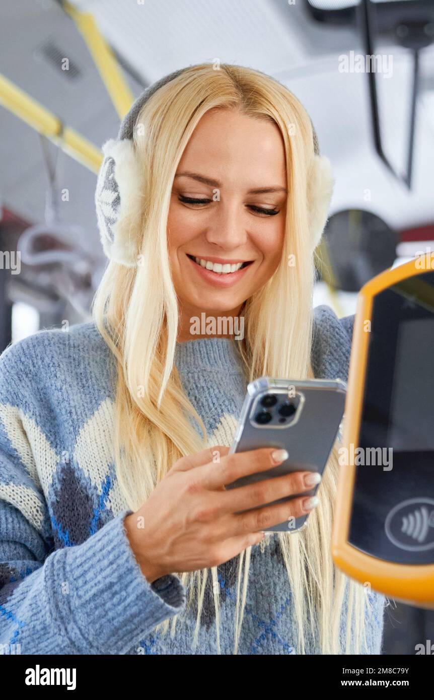 Portrait of happy smiling fair long haired with warm earmuffs female staring in phone holding it near the terminal paying fare by Paypass. Beggining of new working day. Concept of morning routine.  Stock Photo