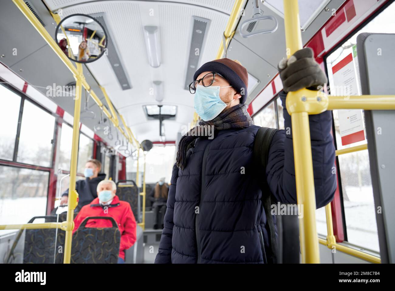 Portrait of young standing man with glasses wearing warm black and brown hat gloves and scarf with coat and two travellers protecting themselves during pandemic with respirator getting work by bus.  Stock Photo