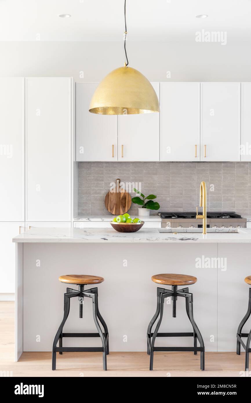 A kitchen detail with white cabinets, gold faucet and light hanging over the island with bar stools, and a tiled backsplash. Stock Photo