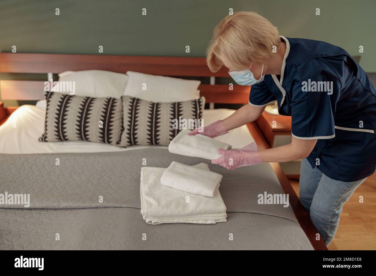 Housemaid in mask putting stack of fresh white bath towels on bed sheet